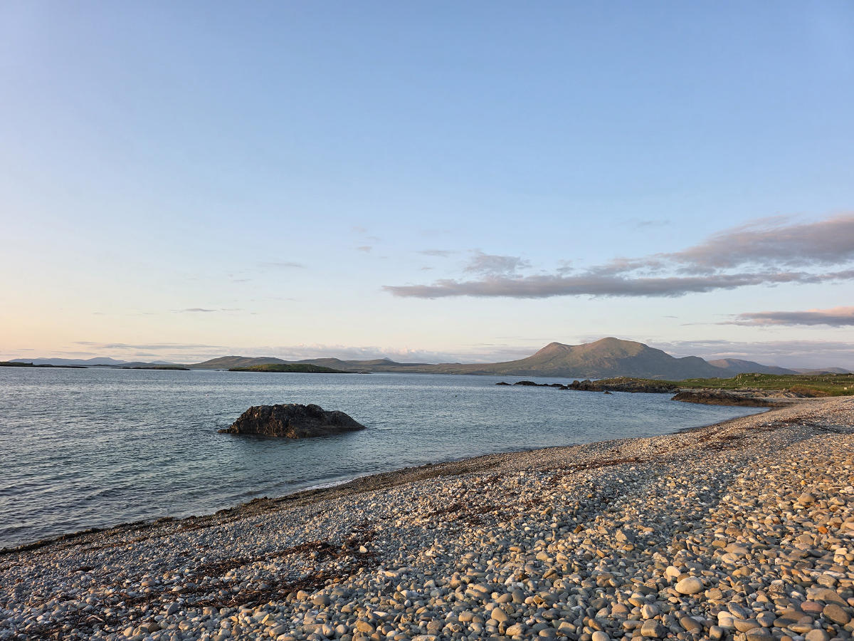 Eine felsige Küste mit ruhigem Wasser, ein großer Felsen in Küstennähe, weit entfernte grüne Inseln und Berge unter einem klaren Himmel bei Sonnenuntergang - perfekte Aussichten in der Nähe von Spitzenhotels in Connemara Co Galway, wie dem renommierten Renvyle House Hotel & Resort.