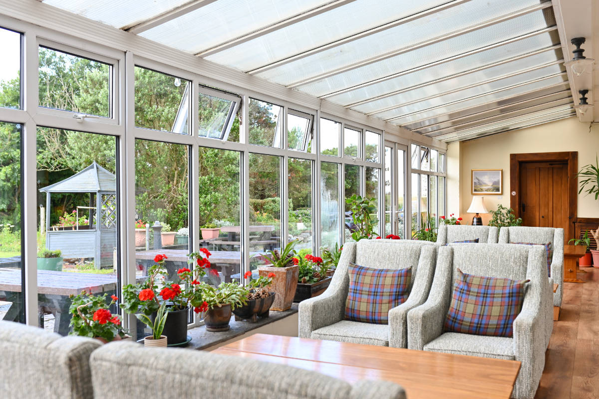 Der helle Sonnenraum im Renvyle House verfügt über große Fenster, Sessel, einen Holztisch, rote Topfblumen und Blick auf einen Garten mit Pavillon - ein ruhiger Rückzugsort in einem der besten 4-Sterne-Hotels Connemaras.