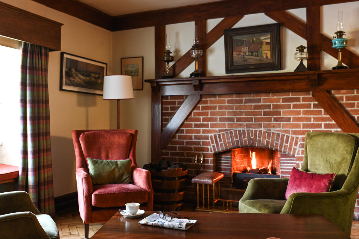 Das gemütliche Wohnzimmer im Renvyle House Hotel & Resort ist mit einem beleuchteten Ziegelsteinkamin, roten und grünen Sesseln, einem Couchtisch mit einer Tasse und einer Zeitung sowie gerahmter Kunst ausgestattet - perfekt zum Entspannen in einem der besten Hotels, die Irland zu bieten hat.