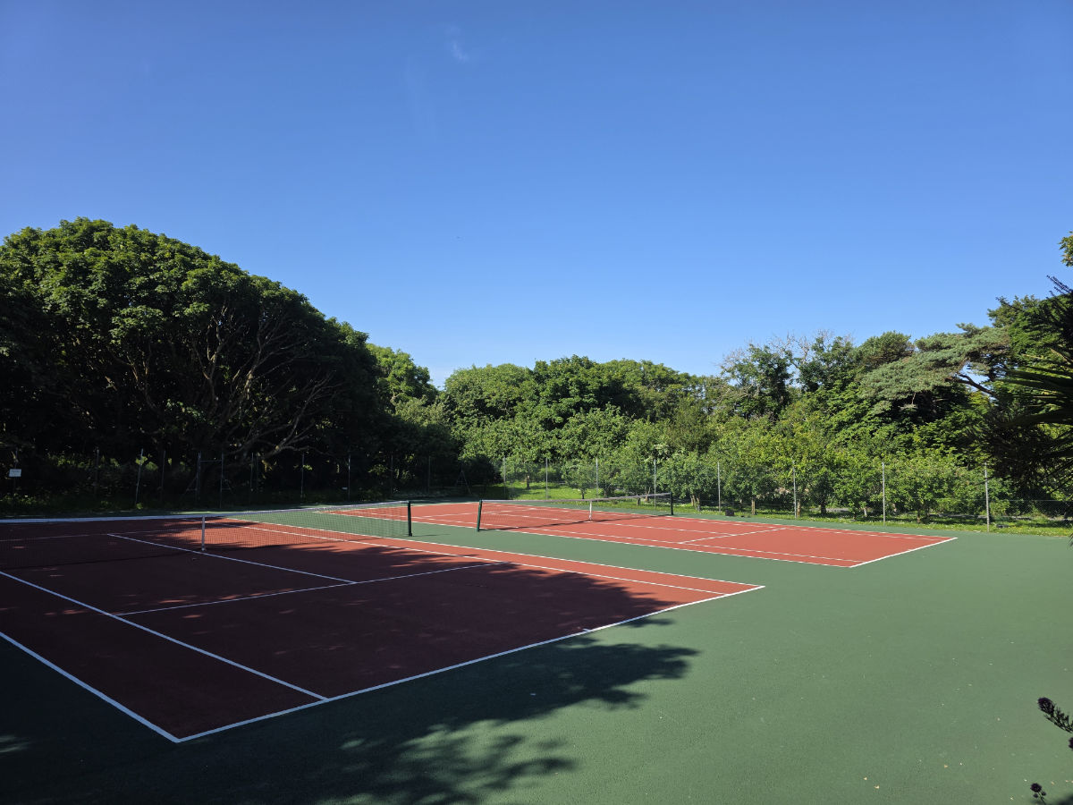 Zwei leere Tennisplätze im Freien mit rotem Belag und grüner Umgebung liegen unter einem klaren blauen Himmel und bieten einen friedlichen Rückzugsort in der Nähe einiger der besten Hotels, die Irland zu bieten hat, darunter 4-Sterne-Hotels in Connemara.