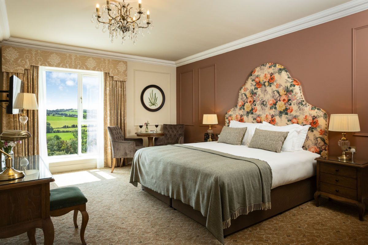 Elegante camera d'albergo al Fernhill House Hotel con testata floreale, letto king, due comodini, tavolino con sedie, lampadario e finestra con vista sulla rigogliosa campagna irlandese. Perfetta per la vostra fuga in un hotel di campagna in Irlanda.