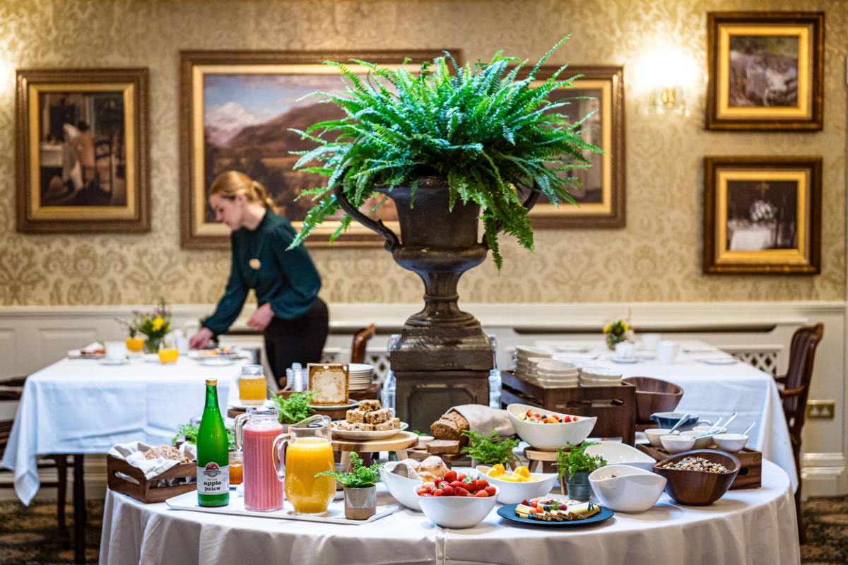 Un tavolo da buffet con vari cibi e bevande per la colazione è allestito in una sala da pranzo decorata del Fernhill House Hotel; una donna sistema gli oggetti su un tavolo sullo sfondo di questo affascinante hotel a 4 stelle in Irlanda.