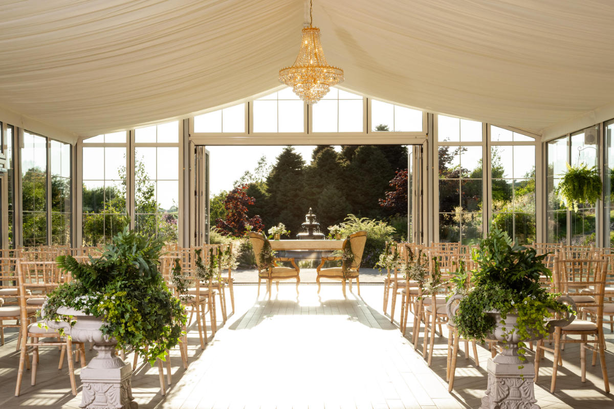 L'allestimento di una cerimonia nuziale al Fernhill House Hotel è caratterizzato da file di sedie, verde lussureggiante, due sedie decorate e un tavolo che si affaccia su un giardino aperto sotto un lampadario in questo affascinante hotel a 4 stelle in Irlanda.