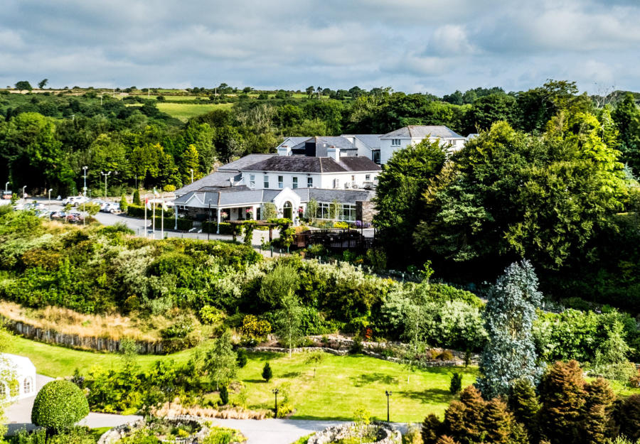Le Fernhill House Hotel est un grand hôtel de campagne en Irlande, entouré d'arbres denses et de jardins paysagers, avec un parking et des champs verts en arrière-plan sous un ciel partiellement nuageux.