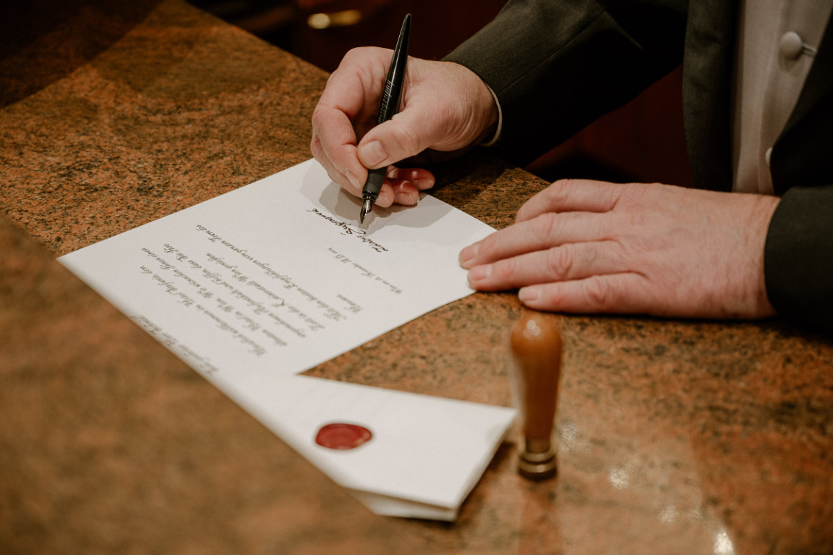 A person signs a document on a brown marble surface at Hotel Stefanie, with a wax-sealed envelope and a wooden stamp nearby.