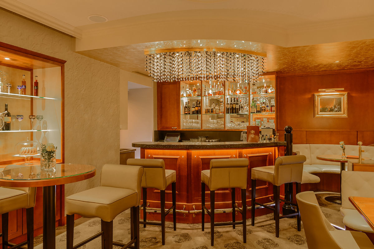 A well-lit, elegant hotel bar at Hotel Stefanie Wien with padded beige chairs, glass tables, a chandelier, and a mirrored back bar stocked with bottles and glasses.