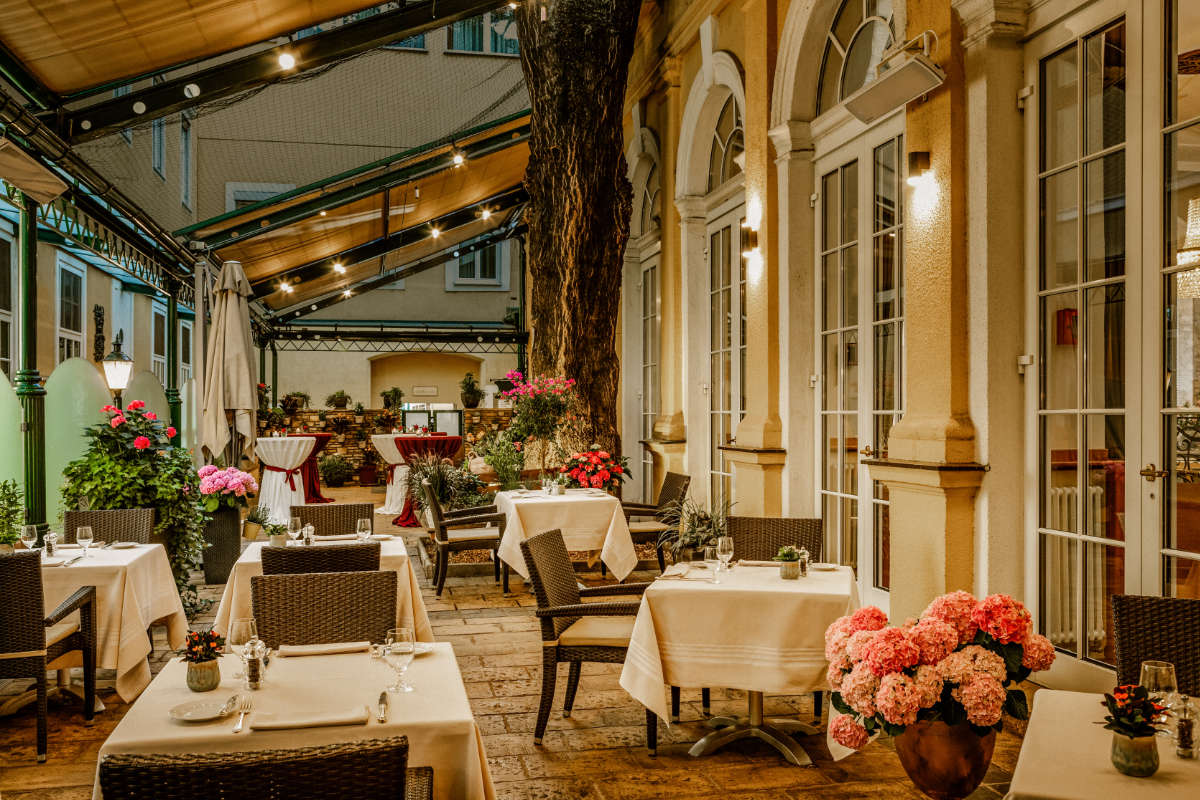 Elegant outdoor restaurant patio at Hotel Stefania Vienna featuring wicker chairs, white tablecloths, potted flowers, and warm lighting next to a building with arched windows—a perfect spot in this 4 star hotel Vienna city centre.