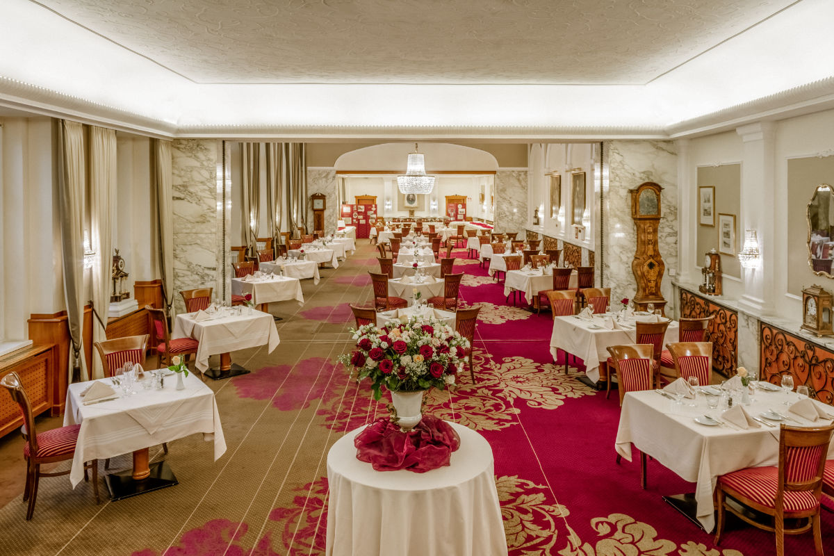 Elegant restaurant dining room at Hotel Stefanie features white tablecloths, red chairs, floral centerpiece, ornate décor, and bright lighting. Tables are set for guests in this classic 4 star hotel Vienna setting with a red carpet and timeless furnishings.