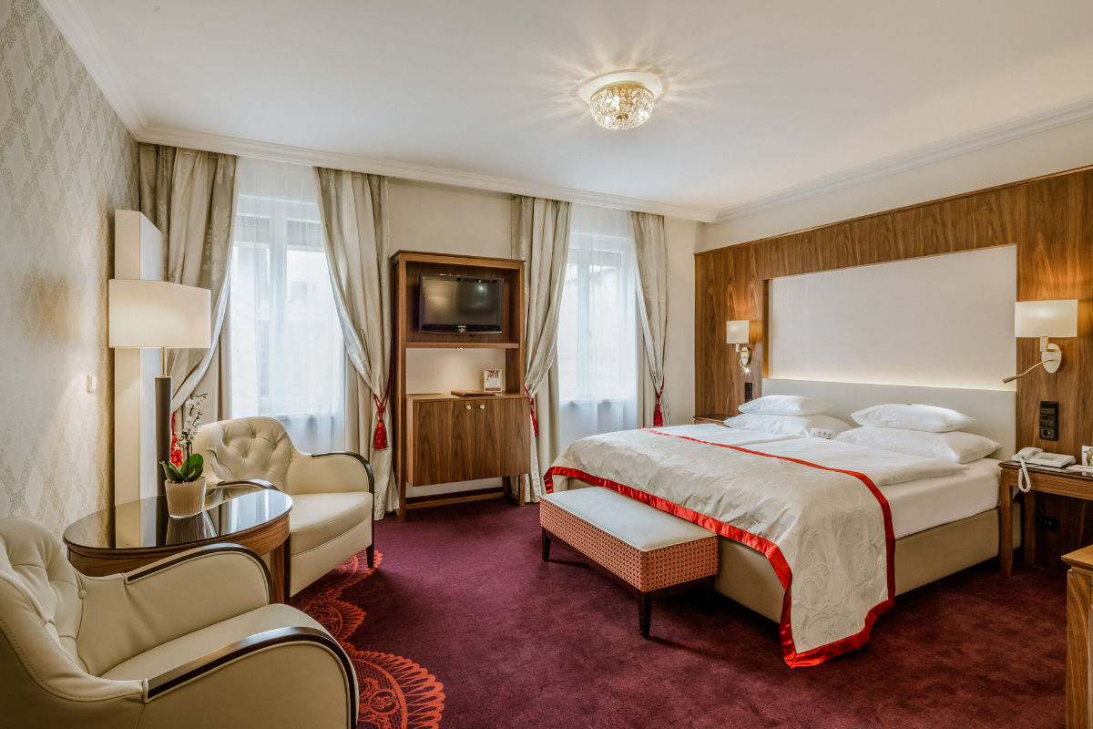 A hotel room at Hotel Stefania Vienna with a double bed, two armchairs, a small round table, wall-mounted TV, and two curtained windows. The decor features beige, white, and red tones—perfect for your 4 star hotel Vienna city centre stay.