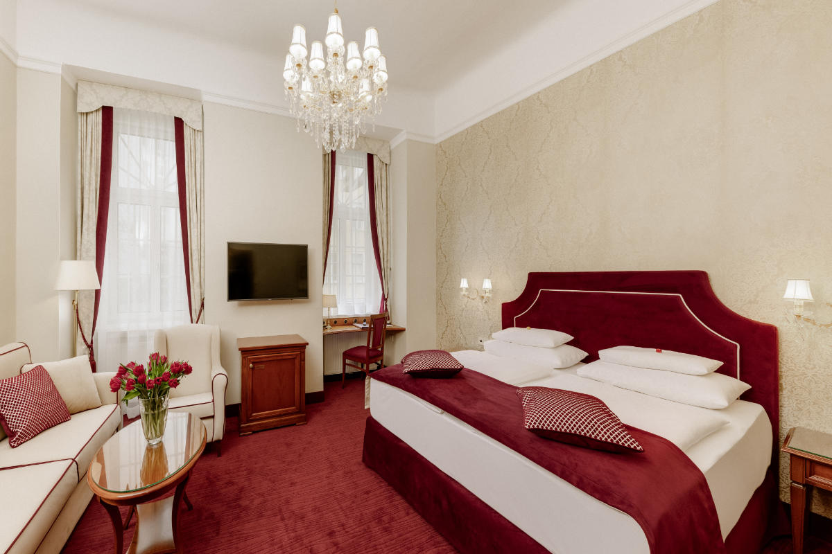 Hotel Stefanie Vienna room featuring a large bed, maroon carpet, chandelier, wall-mounted TV, sofa, small table, desk, and two windows with curtains provides comfort and elegance in the heart of the city.