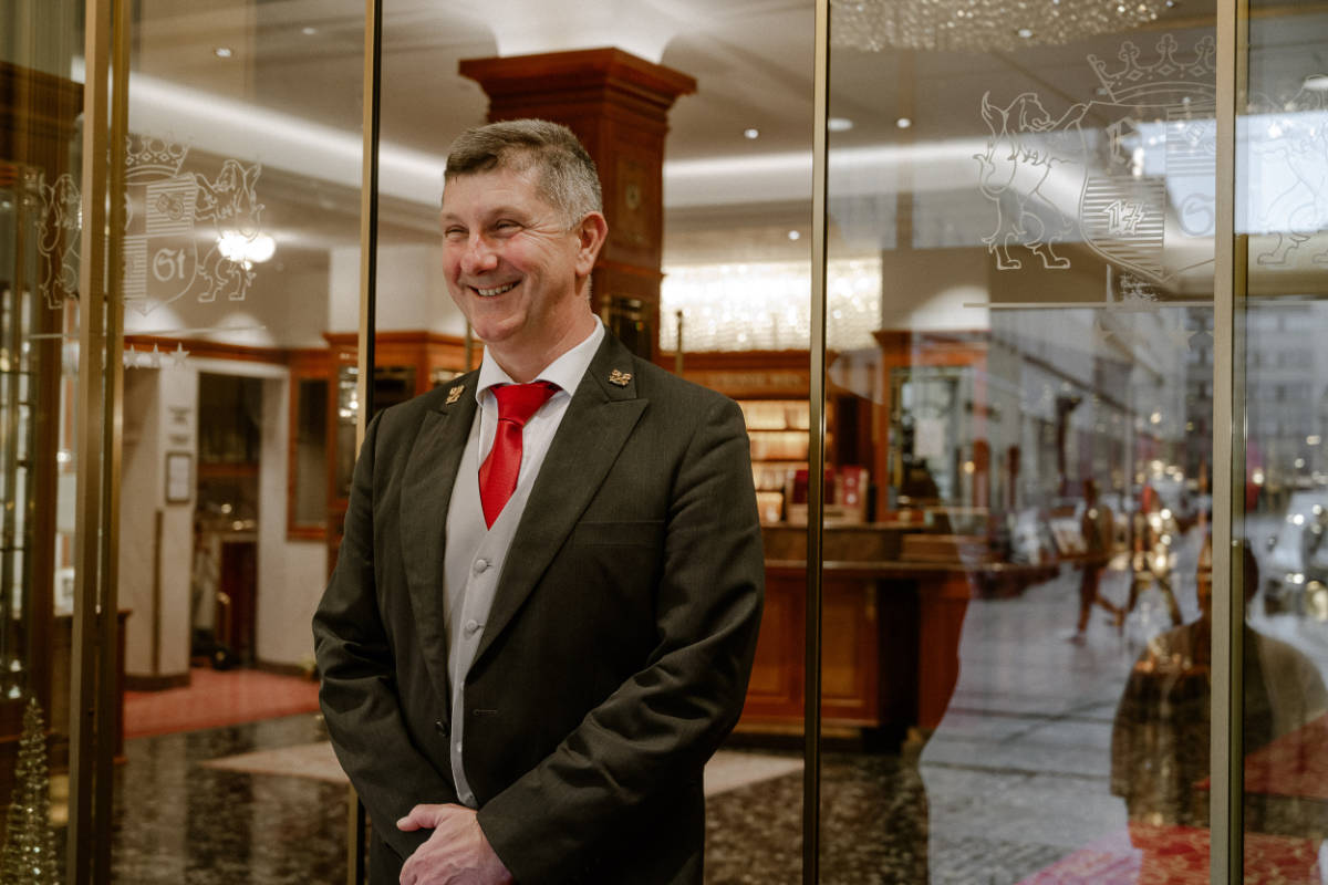 A man in a suit with a red tie stands smiling in front of glass doors at the entrance of an elegant 4 star hotel Vienna city centre lobby.