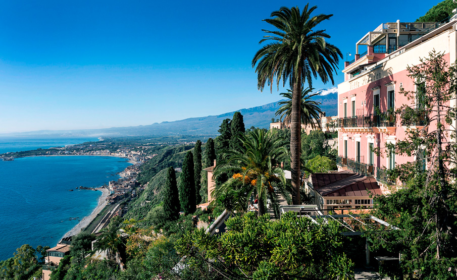 Vue panoramique de la côte avec des bâtiments roses, des palmiers et une végétation luxuriante surplombant un océan bleu et une ville lointaine. Le ciel clair et les collines en arrière-plan encadrent magnifiquement la Villa Schuler Taormina, ajoutant de l'élégance à cet hôtel 4 étoiles pittoresque en Sicile.