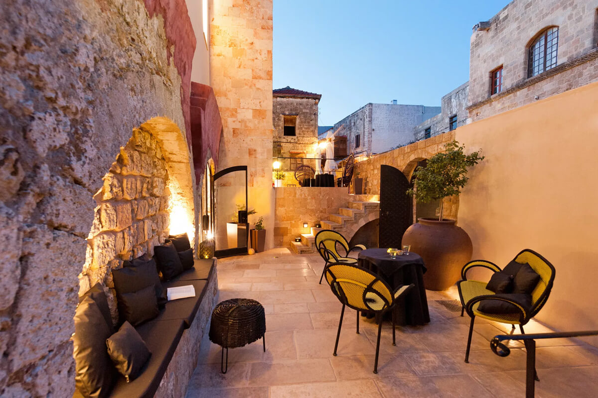 A courtyard with tables and chairs in front of a stone wall at the Allegory Boutique Hotel, Rhodes, Greece.