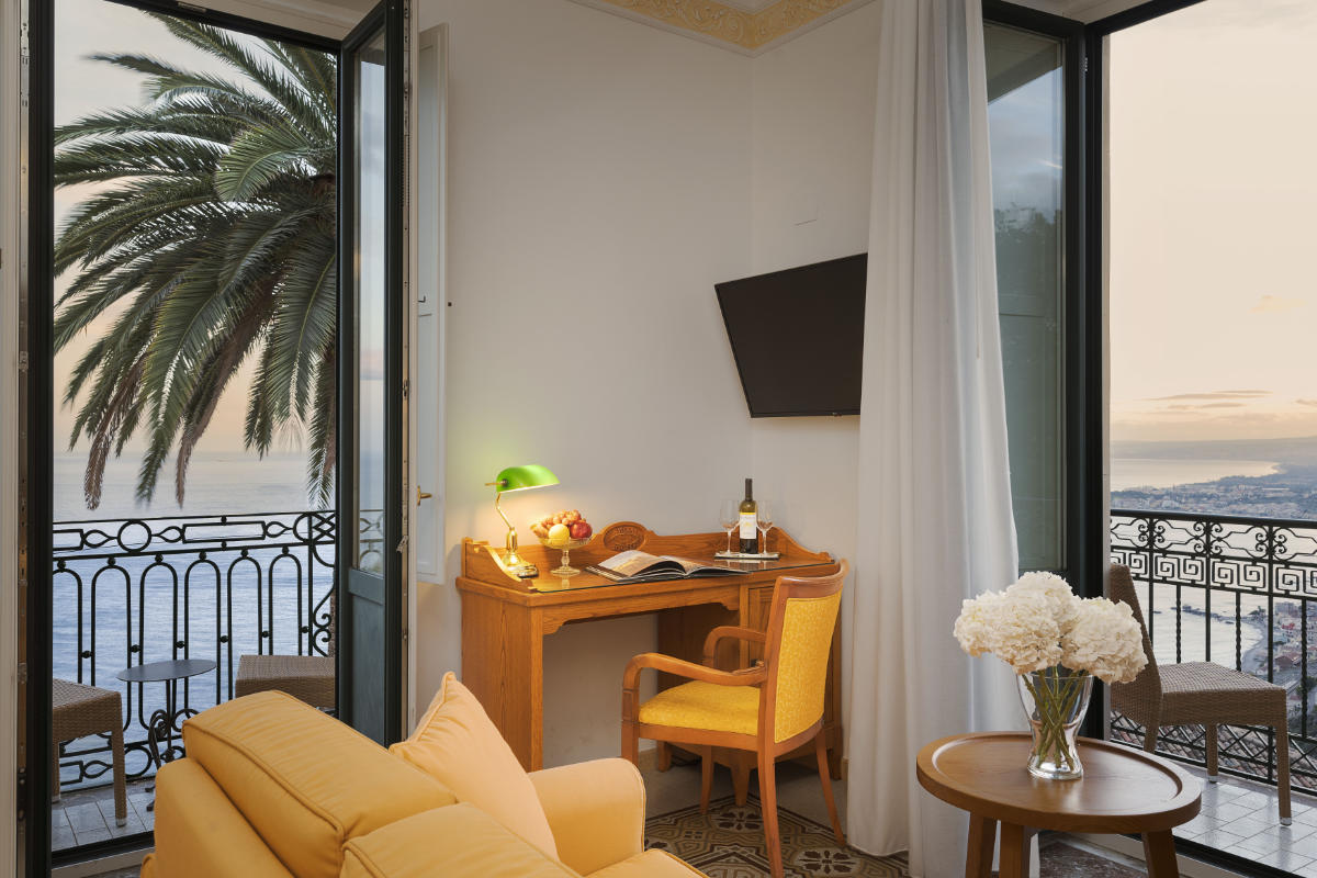 Un'accogliente camera dell'Hotel Villa Schuler si affaccia su un balcone con una palma e il tramonto sull'oceano: l'ideale per rilassarsi in questo incantevole hotel 4 stelle in Sicilia.