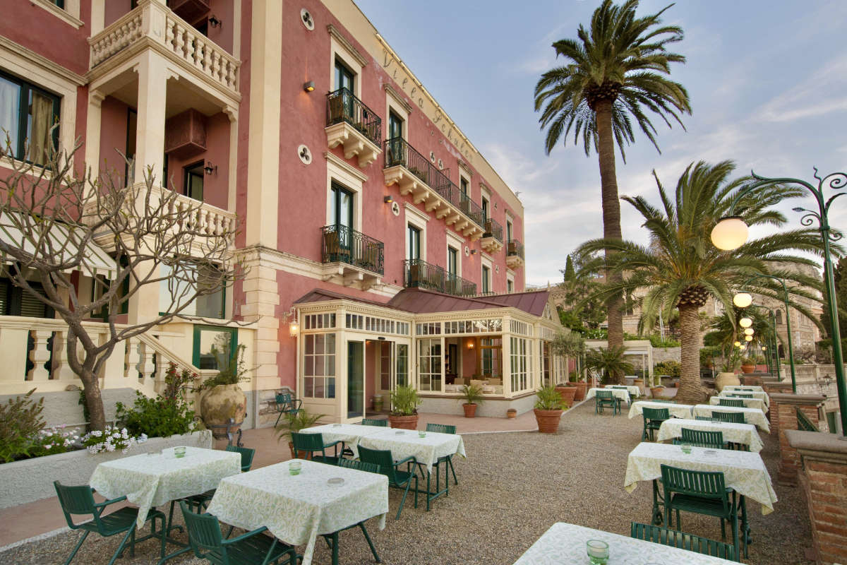 The outdoor patio of Hotel Villa Schuler, a 4-star hotel in Taormina, features tables and chairs set against the pink facade of the building, surrounded by lush palm trees and greenery.