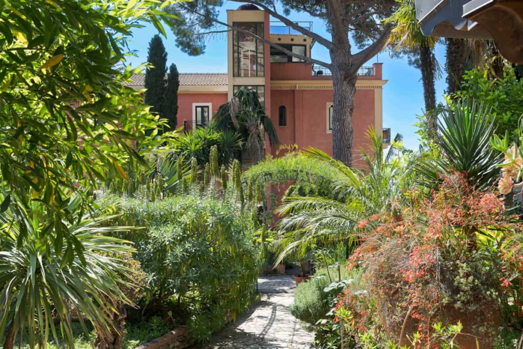 L'éveil du printemps : Les jardins européens prennent vie dans le parc d'un hôtel historique 1 Un chemin de pierre serpente à travers un jardin verdoyant et luxuriant vers l'hôtel Villa Schuler, un hôtel 4 étoiles en Sicile, avec des balcons et de grands arbres sous un ciel bleu clair.