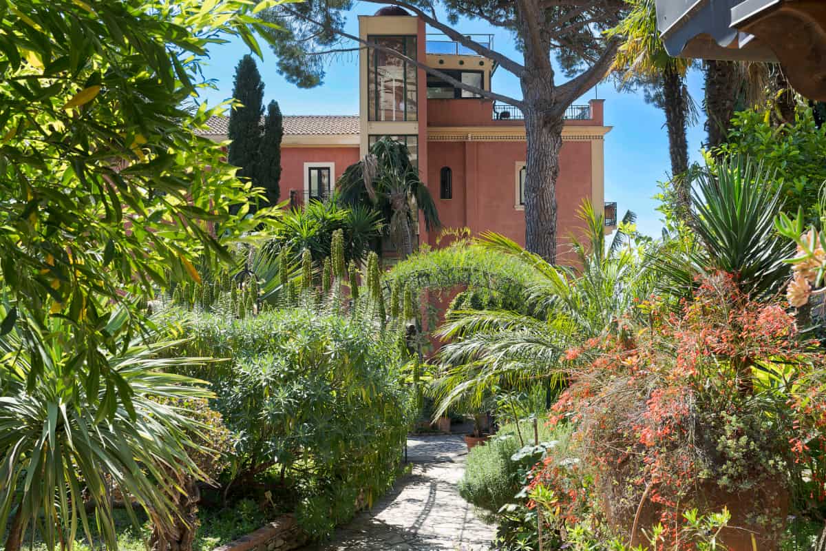 Un sentiero di pietra si snoda attraverso un giardino verde e lussureggiante verso l'Hotel Villa Schuler, un albergo a 4 stelle in Sicilia, color terracotta, con balconi e alberi ad alto fusto sotto un cielo azzurro e limpido.