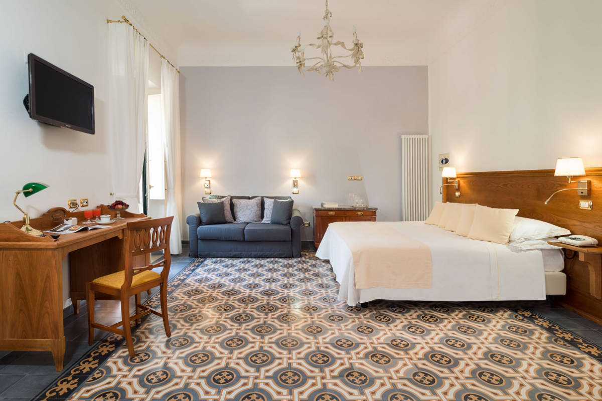 Experience the elegance of a Hotel Villa Schuler room, featuring a double bed, sofa, desk, and TV. The spacious room boasts decorative floor tiles and a chandelier. Discover luxury at this 4-star hotel in Sicily.