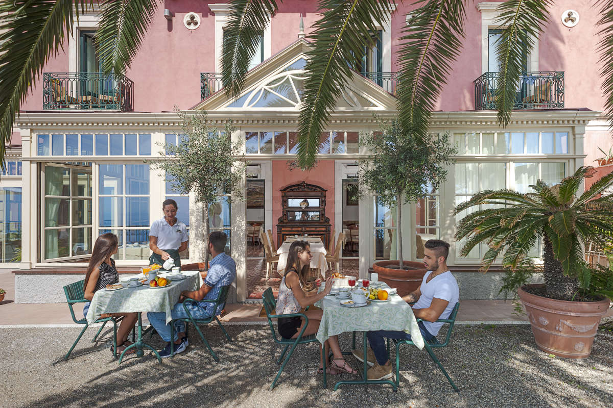 People sitting at outdoor tables enjoying breakfast under palm trees in front of a large pink building with many windows at Villa Schuler Taormina, a charming 4-star hotel in the heart of Sicily.
