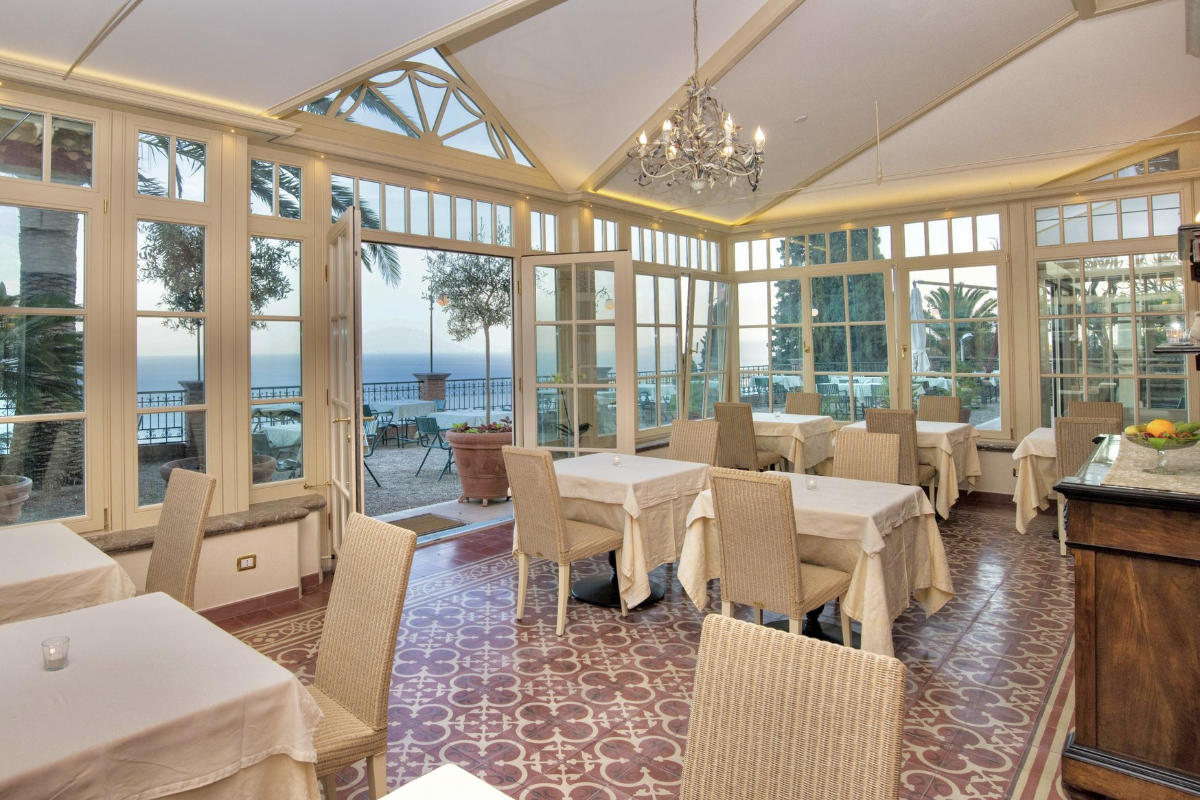 The bright conservatory dining area at Hotel Villa Schuler Taormina boasts large windows, a tiled floor, and tables adorned with white cloths. Enjoy a stunning view of the outdoor terrace and sea as a chandelier elegantly hangs from the ceiling.