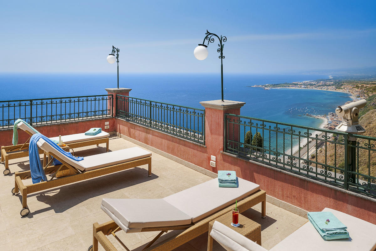 The rooftop terrace at Hotel Villa Schuler features lounge chairs and a telescope, offering a coastal view of the ocean and distant shoreline under a clear blue sky, capturing the essence of Taormina's scenic beauty.