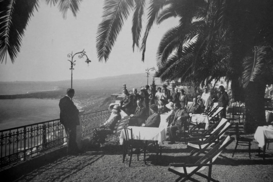 A group of people gathers on a terrace with tables and chairs under palm trees, overlooking the breathtaking coastal view at Hotel Villa Schuler in Taormina, Sicily.