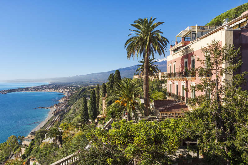 Il Club del libro degli alberghi storici: Fughe letterarie attraverso l'Europa 8 Vista costiera di una città mediterranea con verde lussureggiante, un edificio rosa e palme vicino al rinomato Hotel Villa Schuler a Taormina, in Sicilia. Il litorale lontano scintilla sotto un cielo azzurro e limpido.