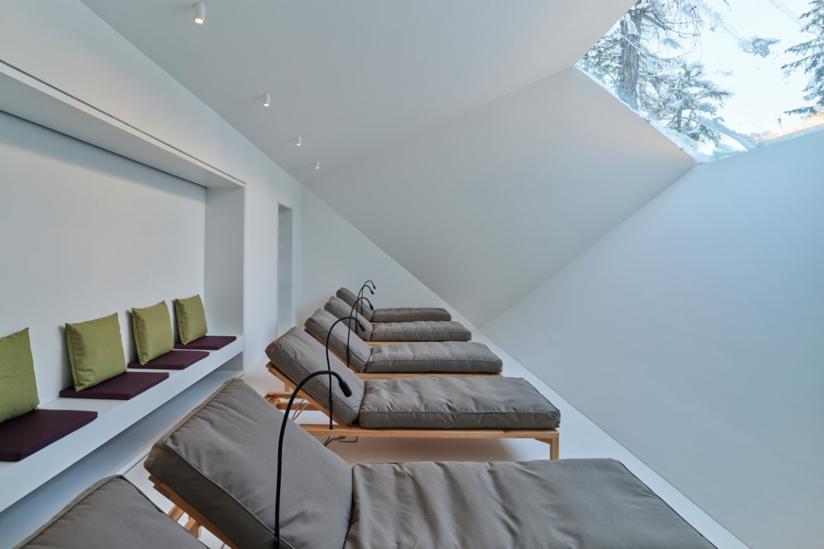 Eine Reihe von gepolsterten Lounge-Sesseln mit Leselampen in einem modernen, minimalistischen Zimmer im Hotel Waldhaus Sils - einem renommierten historischen Hotel in der Schweiz - zeichnet sich durch große, schräge Fenster und grüne Akzentkissen aus.