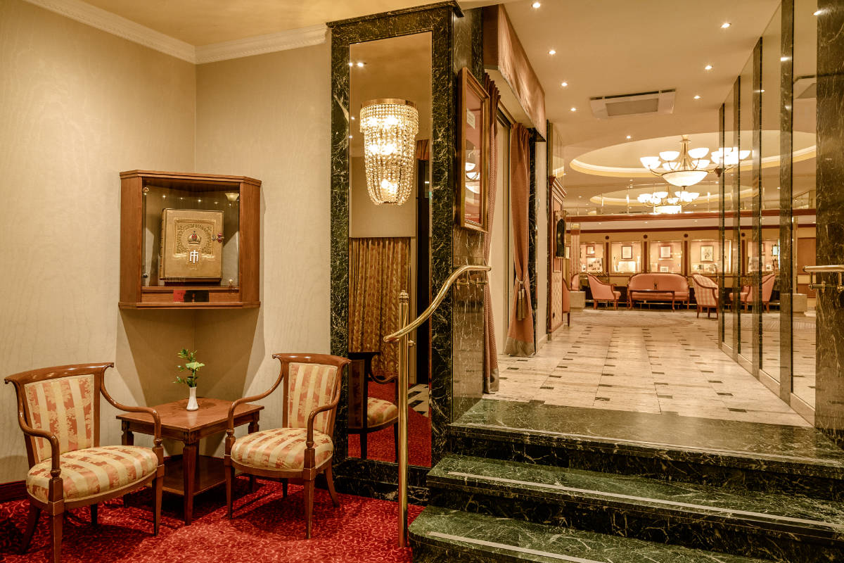 L'elegante hall dell'Hotel Erzherzog Rainer è caratterizzata da gradini in marmo verde, illuminazione con lampadari e sedute eleganti. Due sedie e un tavolino sono perfettamente posizionati vicino a un'esposizione in vetro, mentre il pavimento passa senza soluzione di continuità dalla moquette al marmo.