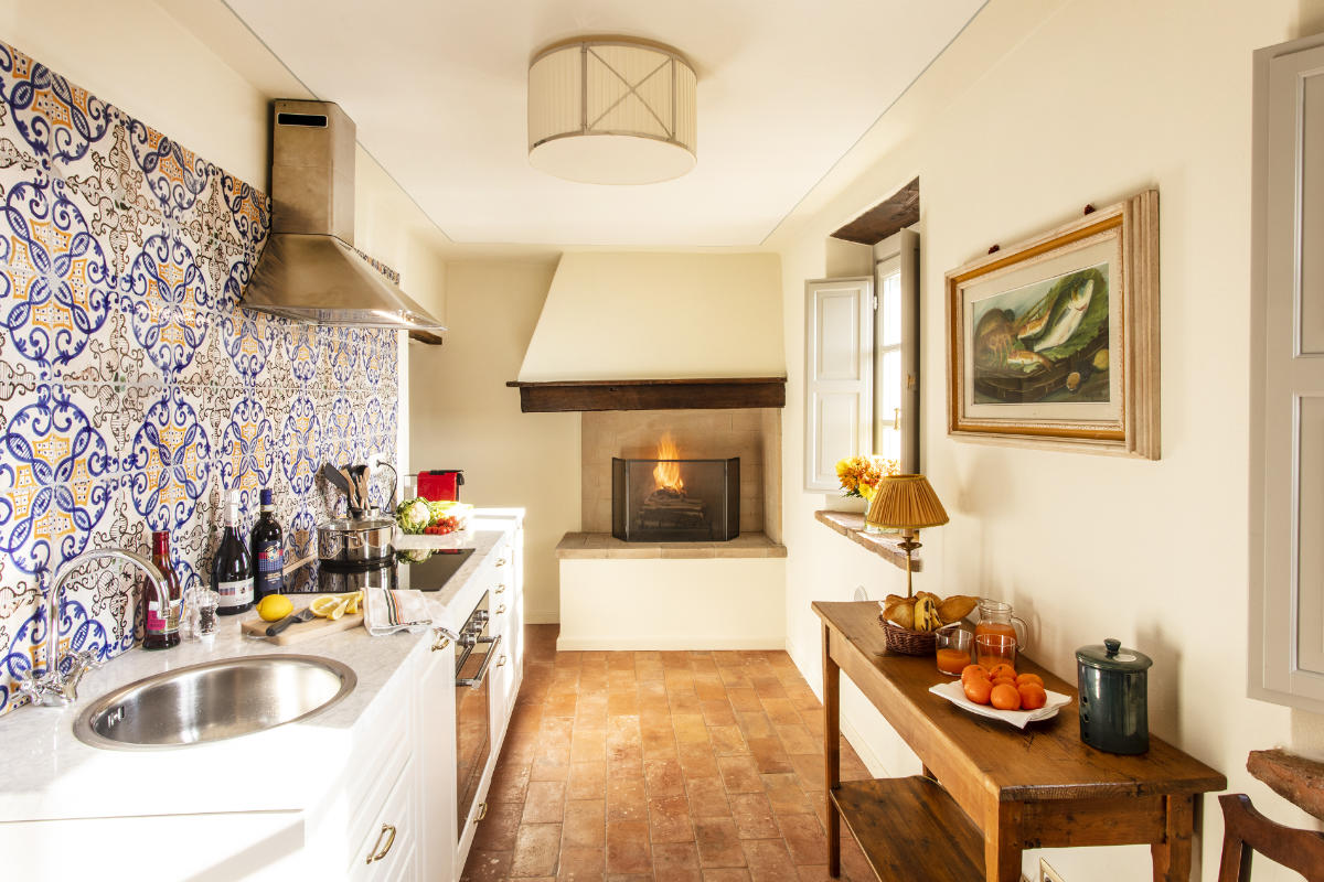 Una acogedora cocina que recuerda a los hoteles de lujo de la Toscana cuenta con un panel de azulejos estampados, electrodomésticos de acero inoxidable, una chimenea y una mesa de madera con fruta y una lámpara. En la pared cuelga un cuadro que deja entrever el encanto propio del Palazzo Tiglio.