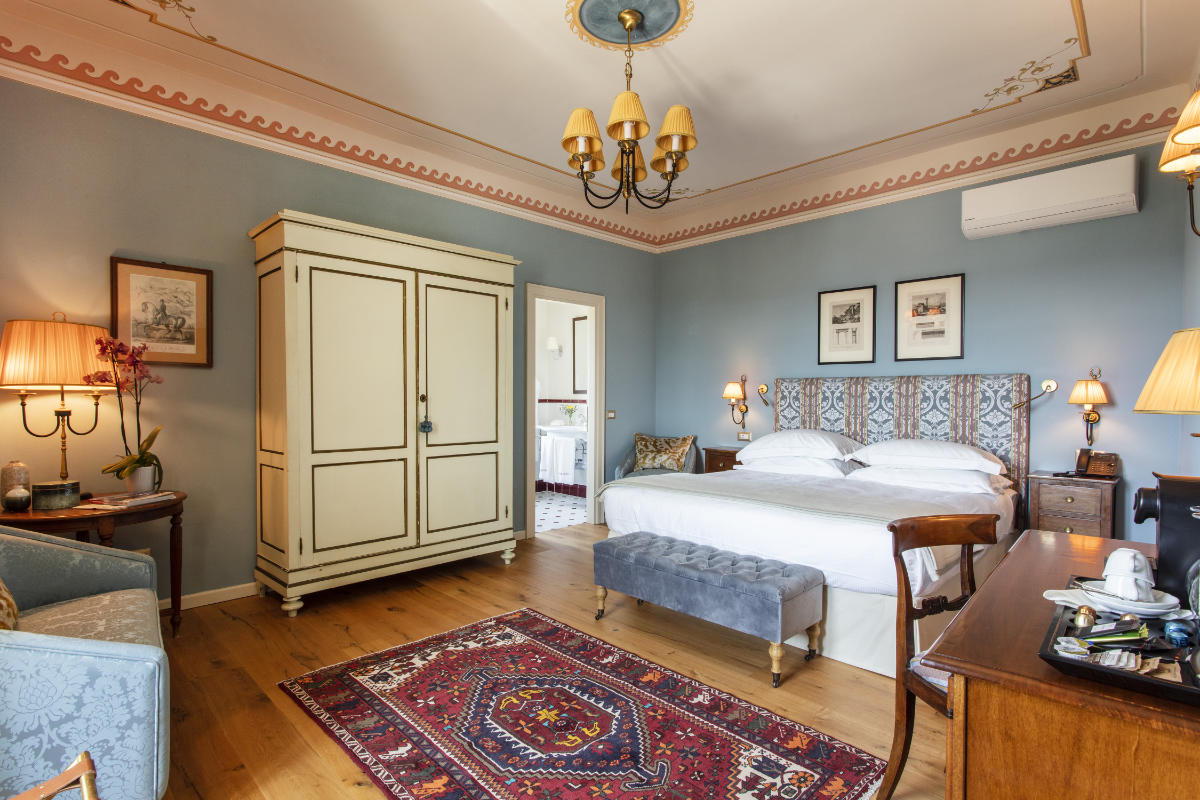 Un dormitorio bien decorado con paredes azules, que recuerda a una suite de los hoteles de lujo de la Toscana, cuenta con una cama, un armario, un escritorio de madera, sillas y una alfombra ornamentada. Del techo cuelga una lámpara de araña. Lámparas y cuadros completan la decoración de este sereno refugio.
