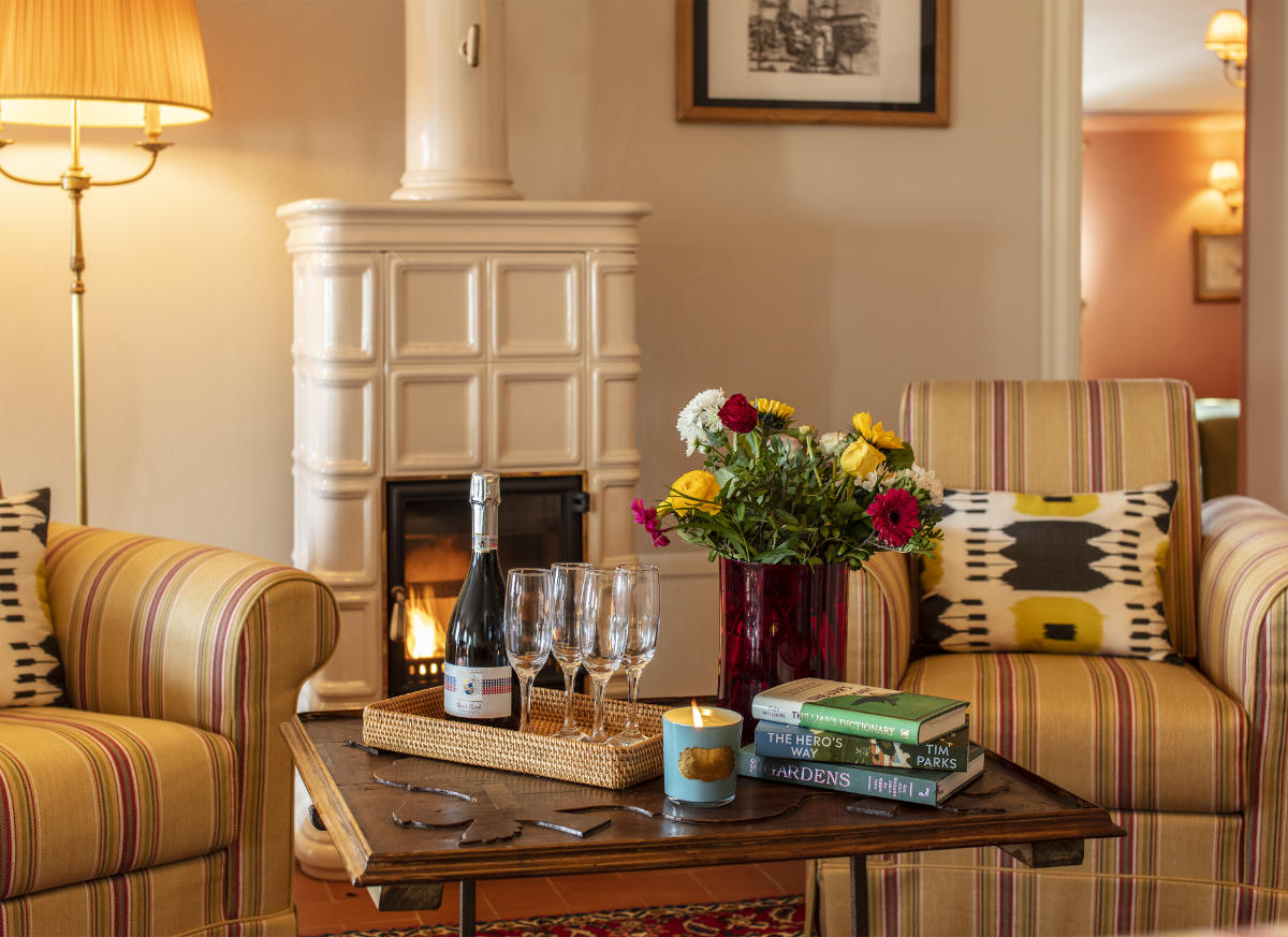 Una acogedora sala de estar que recuerda a los hoteles de lujo de la Toscana, con una mesa con una botella de vino, copas y libros. Dos sillones a rayas descansan junto a un arreglo floral y una lámpara, y una chimenea de cerámica blanca completa esta escena que evoca el encanto del Palazzo Tiglio.