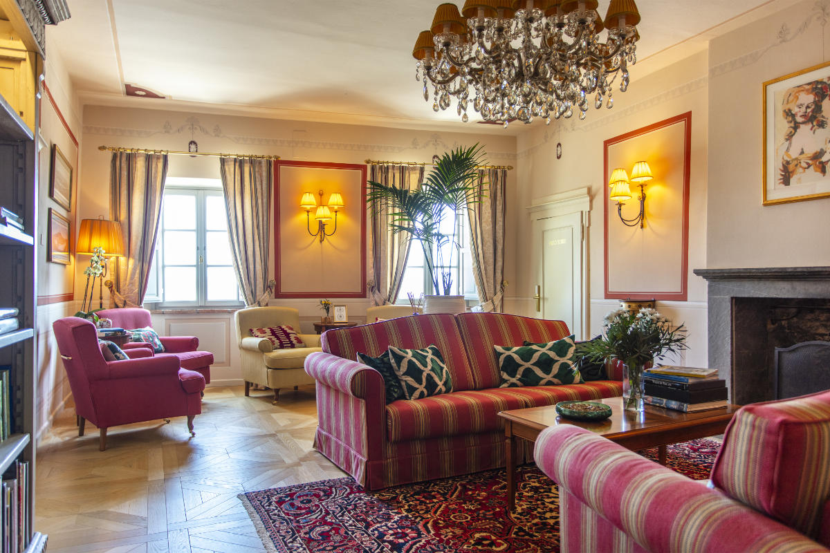 Una acogedora sala de estar que recuerda a los hoteles de lujo de la Toscana, con sofás a rayas, sillones, una lámpara de araña y una alfombra decorativa. Una planta junto a la ventana y libros sobre la mesa contribuyen al ambiente. Las paredes están decoradas con obras de arte e iluminación similar a la del Palazzo Tiglio.