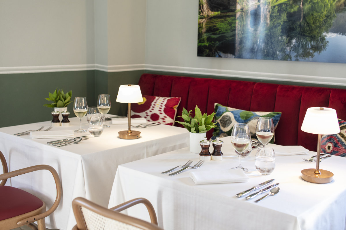 Dos mesas de comedor elegantemente dispuestas con manteles blancos, copas de vino, lámparas de mesa y plantas en macetas evocan el encanto de la Toscana. Los asientos acolchados en rojo y las paredes verdes adornadas con obras de arte de la naturaleza crean un ambiente que recuerda al Palazzo Tiglio de los hoteles de lujo de la Toscana.