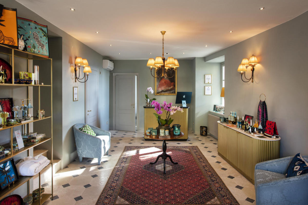 Un acogedor interior de boutique que recuerda a los hoteles de lujo de la Toscana, con una elegante decoración que incluye una mesa central adornada con orquídeas, una alfombra con motivos geométricos y una iluminación ambiental de lámparas de araña y apliques de pared. En las estanterías de pared se exponen productos selectos que reflejan el encanto del Palazzo Tiglio.