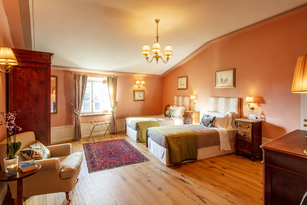 Un dormitorio cálidamente iluminado al estilo de los hoteles de lujo de la Toscana, con dos camas, suelo de madera, sillón y ventana. Las paredes rosas se complementan con una alfombra chic y varias lámparas y muebles que recuerdan el encanto del Palazzo Tiglio.