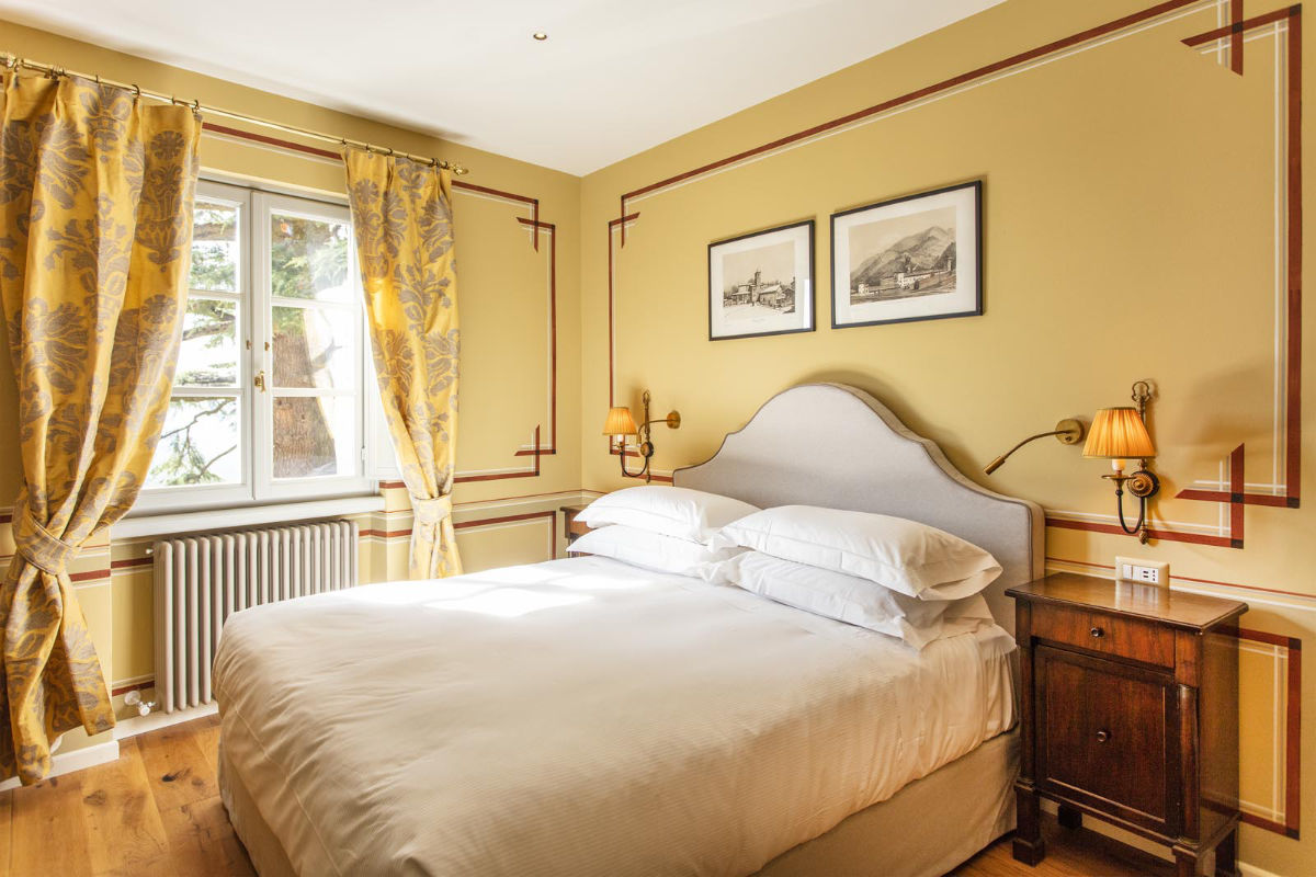 Un dormitorio ordenado que recuerda a los hoteles de lujo de la Toscana, con una cama doble, paredes amarillas, cortinas estampadas, dos lámparas de pared, cuadros enmarcados y una mesilla de noche de madera cerca de una ventana que evoca la elegancia del Palazzo Tiglio.
