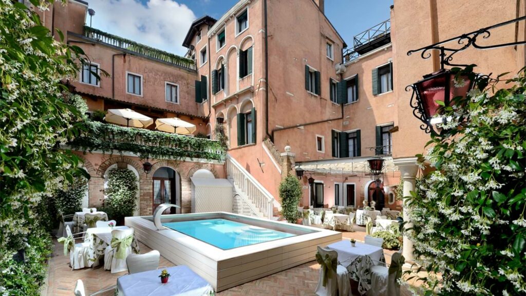 Capodanno negli hotel storici d'Europa 10 Cortile esterno dell'Hotel Giorgione a Venezia, Italia, con una piccola piscina rettangolare circondata da tavoli e sedie, vegetazione lussureggiante e pareti color pesca con balconi e finestre.