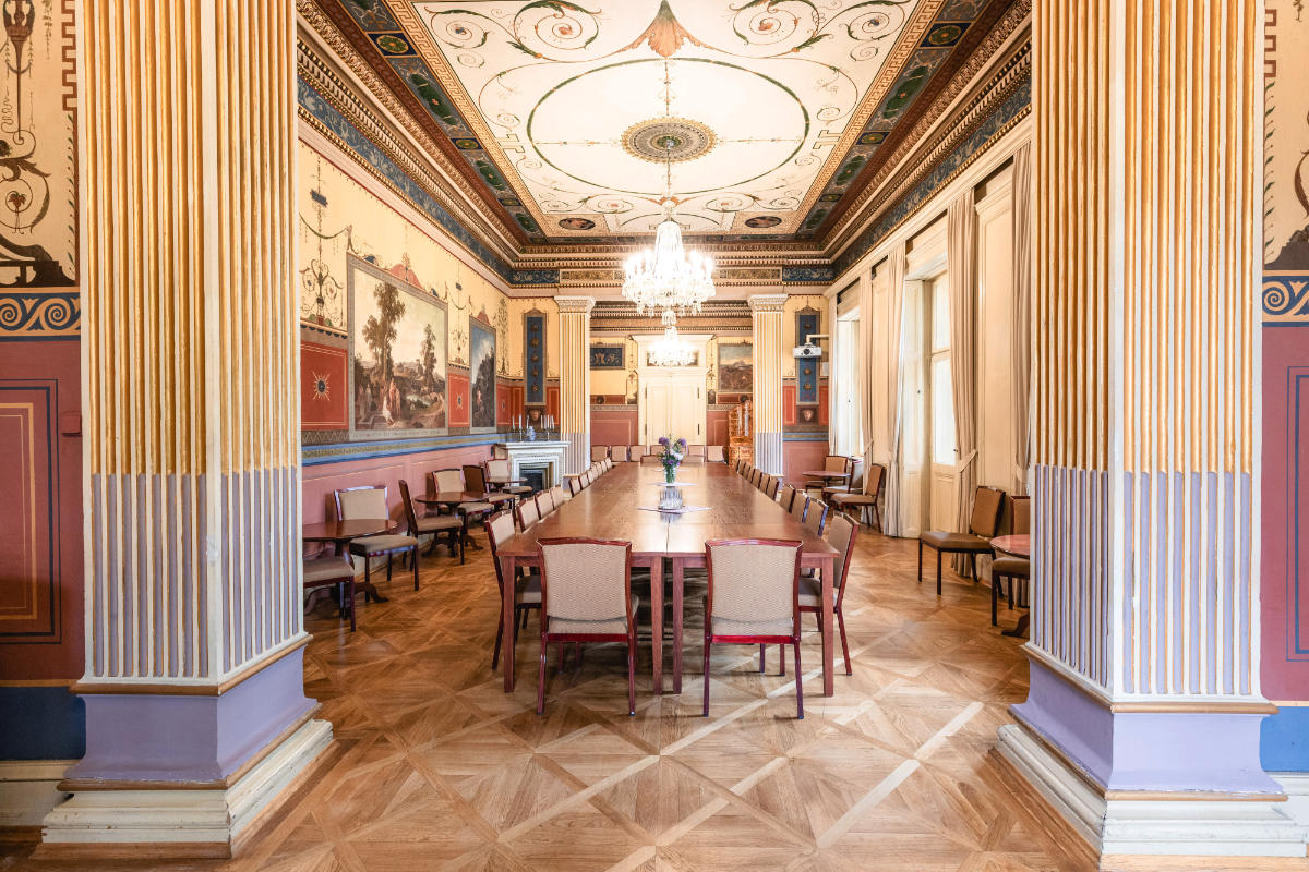 Una larga mesa rectangular con sillas ocupa la ornamentada sala de reuniones Villa Lanna de Praga, con techos altos, detalladas obras de arte en paredes y techos, lámparas de araña y grandes ventanales.
