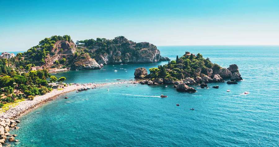A picturesque coastal landscape with a clear blue sea, a pebbled beach, and rocky islets near a verdant headland offers many things to do in Europe over Easter.