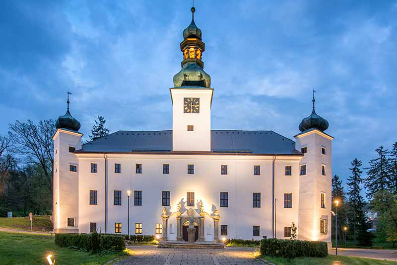 Château baroque illuminé au crépuscule avec des statues ornées devant, entouré de verdure luxuriante, sous un ciel nuageux.