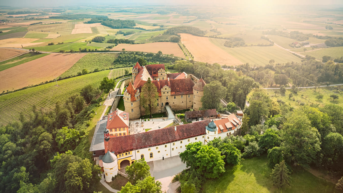 Vista aérea de Schloss Frankenberg, un gran castillo histórico rodeado de exuberante vegetación y campos en un día soleado.