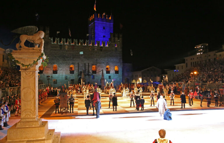 A medieval festival in Marostica, Italy, at night in front of a castle, featuring performers in costumes, musicians, and chess games.