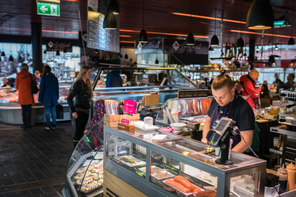 A worker arranging items at a deli counter in a bustling indoor market, featured in the city guide for Bergen.