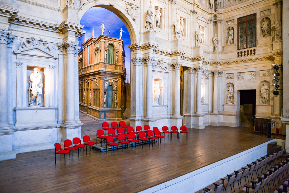 L'ornato palcoscenico del teatro, che ricorda le Meraviglie architettoniche del Palladio, è caratterizzato da statue, colonne bianche e un fondale ad arco. File di sedie rosse sono meticolosamente disposte sul pavimento, creando un'ambientazione che ricorda i capolavori vicentini di Andrea Palladio riconosciuti dall'UNESCO Italia.