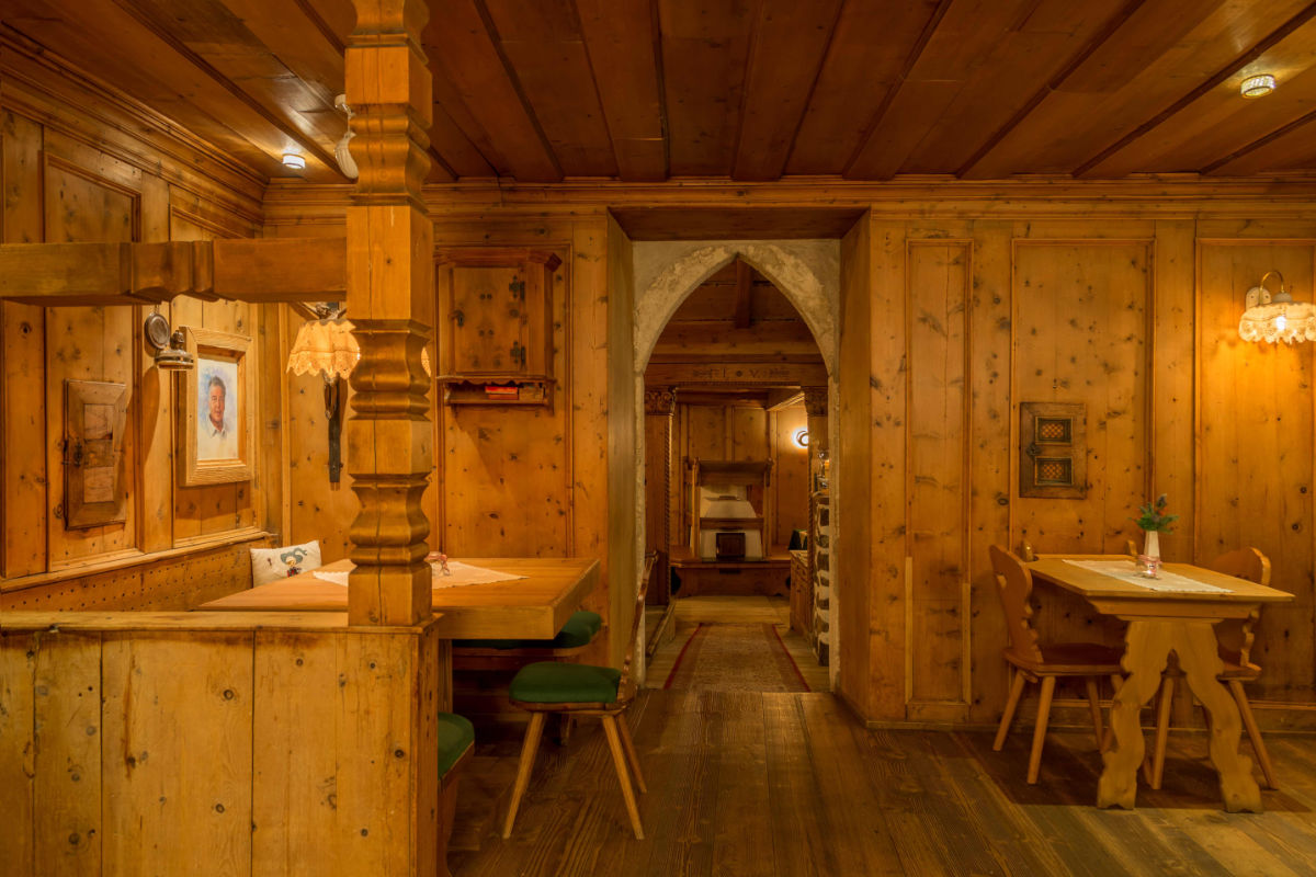 Ein gemütlicher, rustikaler Raum mit Holzwänden und -möbeln, dekoriert mit gerahmten Bildern und kleinen Lampen. Es gibt einen Tisch mit Stühlen und eine Sitzbank auf der einen Seite und einen Torbogen, der zu einem anderen Bereich führt.