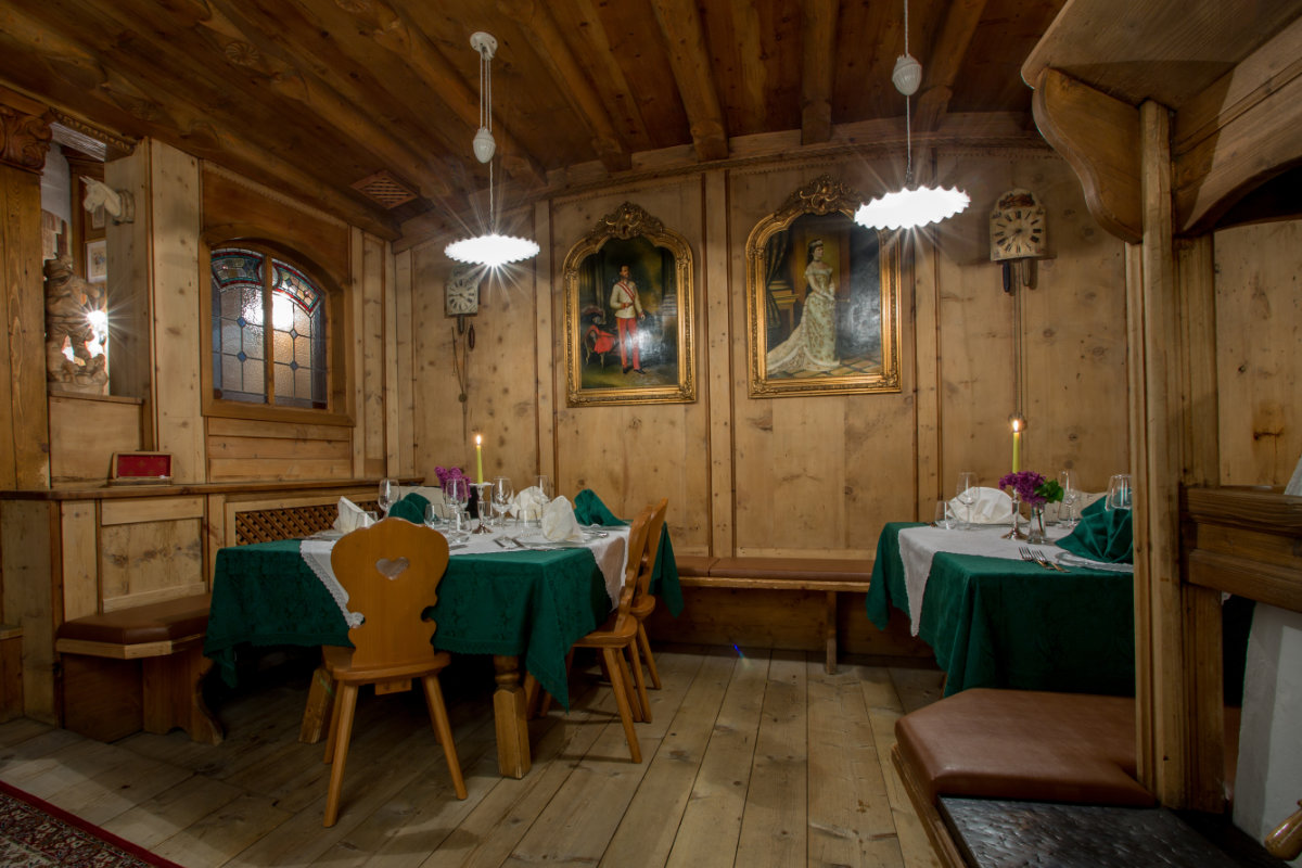 Ein gemütliches Restaurant mit Holzwänden und -decken, dekoriert mit gerahmten Bildern. Es gibt zwei Tische mit grünen Tischtüchern, die ordentlich eingedeckt sind, und Holzstühle. Oberlichter erhellen die Szene.