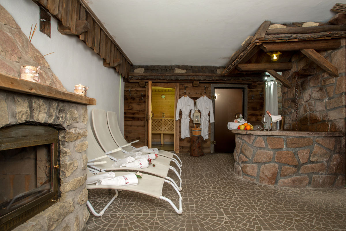 Spa-Raum mit Steinwänden, Liegestühlen, gerollten Handtüchern und Bademänteln, die an den Türen hängen. Ein Obstkorb und Kerzen stehen auf einer steinernen Arbeitsplatte.