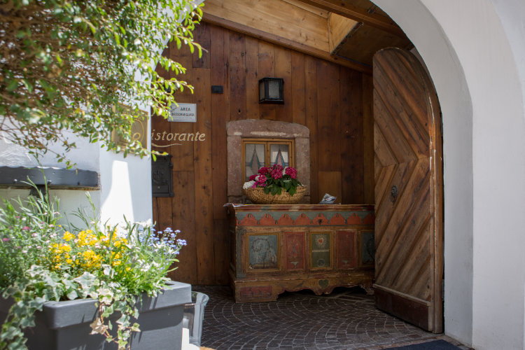 Eine gewölbte Holztür führt in ein rustikales Lokal mit einer mit Blumen geschmückten Holztruhe. Draußen steht ein Pflanzkübel mit gelben und violetten Blumen, und auf einem Schild steht "Ristorante".