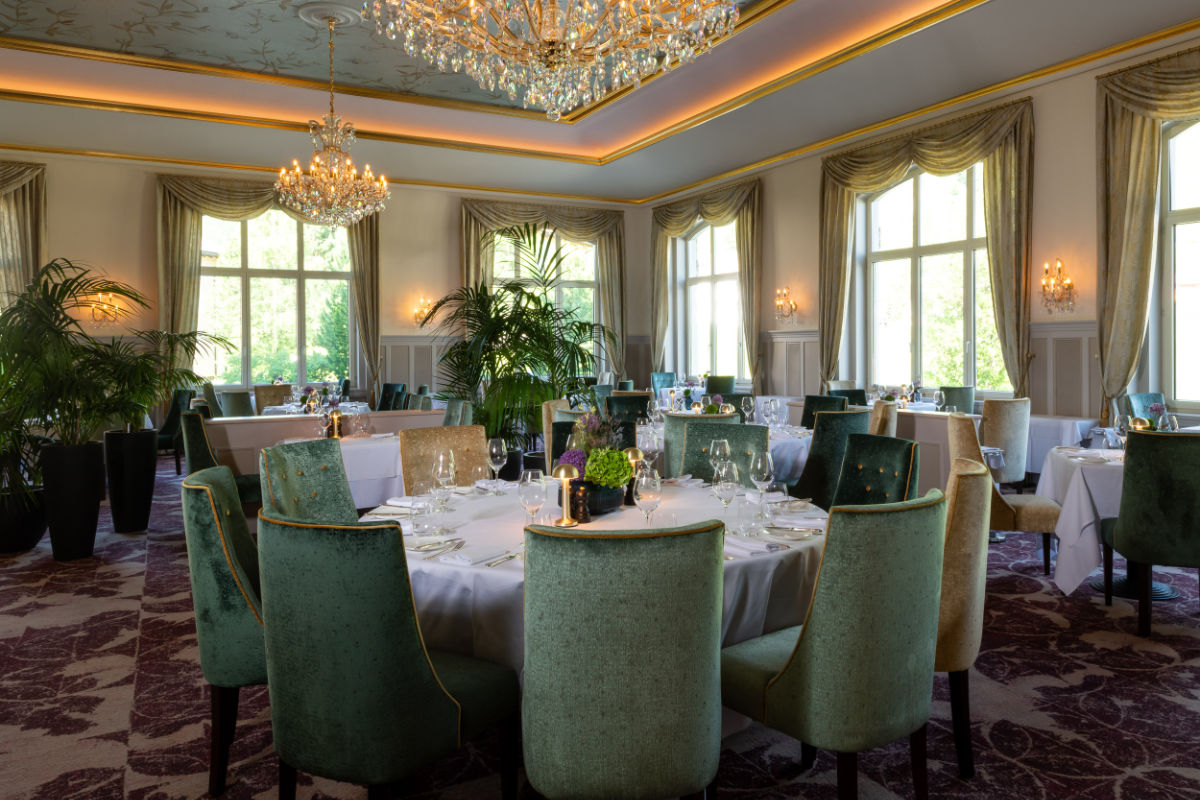 Eleganter Speisesaal im Cresta Palace Hotel Celerina, Schweiz, mit runden Tischen, grün gepolsterten Stühlen, Kronleuchtern, großen Fenstern und Vorhängen - bereit für ein formelles Essen mit edlen Gläsern und Geschirr. Erleben Sie Vier-Sterne-Hotels in der Schweiz.